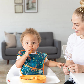 A toddler in a Bumkins SuperBib® eats in a high chair while a smiling woman sits nearby in the cozy living room.