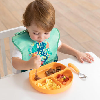 A toddler in a Bumkins SuperBib® enjoys scrambled eggs and fruit with a fork from an orange divided plate at the table.