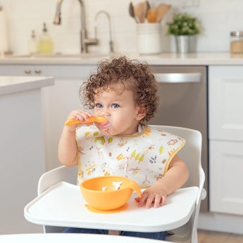 A curly-haired toddler eats with a spoon from an orange bowl, shielded by a colorful Bumkins SuperBib® 3 Pack: Happy Campers.