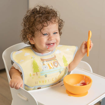 A smiling toddler with curly hair in a high chair holds an orange spoon, wearing the Bumkins SuperBib Happy Camper for easy cleaning.