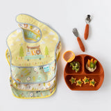 Three Happy Campers SuperBib® by Bumkins, made from waterproof fabric, sit beside a plate and utensil set.