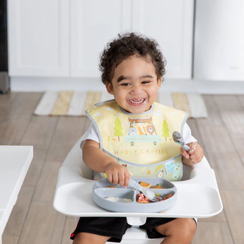 A happy child sits in a highchair, wearing Bumkins SuperBib® Happy Campers, holding a fork, and smiling over a meal in a divided plate.