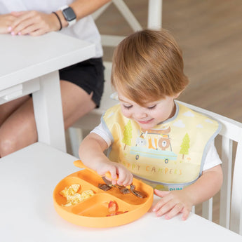 Toddler eats from a sectioned orange plate wearing Bumkins SuperBib® 3 Pack: Happy Campers, sitting at a white table with an adult.