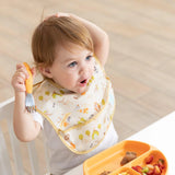 A toddler in a Bumkins SuperBib® 3 Pack: Happy Campers smiles playfully, holding a fork at a table with a mixed food plate.