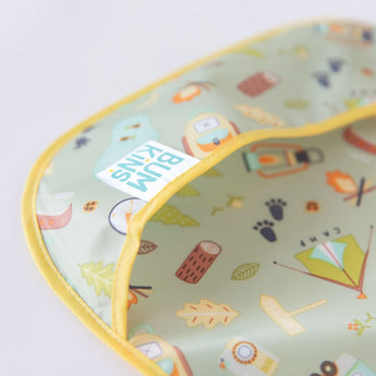 Close-up of Bumkins SuperBib®: Camp Gear with tents, trees, logs & footprints on green. Perfect for your little adventurer!.