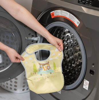 A person washes a Bumkins SuperBib® 3 Pack: Happy Campers (waterproof) in a front-loading machine for easy cleaning.