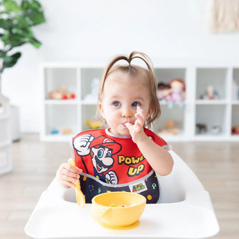 A toddler in a high chair wearing a Bumkins SuperBib®: Super Mario™ Power-Up eats from a yellow bowl, enjoying the easy-wipe fabric.