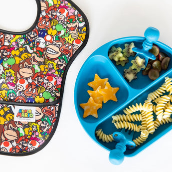 SuperBib® 3 Pack: Super Mario™ Power-Up by Bumkins next to a blue divided plate with cheese stars, pasta, and fruit.