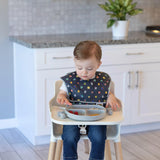 A toddler enjoys a meal from a divided plate, wearing their Bumkins Super Mario™ Power-Up SuperBib® in the kitchen.