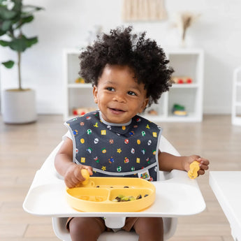 A toddler in a high chair wears a colorful Super Mario™ Power-Up SuperBib by Bumkins, holding a utensil and eating from a yellow plate.