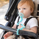 A baby with light brown hair sits in a stroller, wearing a white shirt, exploring with their Bumkins Silicone Tether and pacifier.