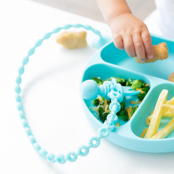 A child reaches for food on a blue plate with nuggets, broccoli, cheese, and fries using the Bumkins Silicone Accessory Tether.