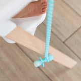 A baby on a swing explores with the Bumkins Silicone Accessory Tether in Blue hanging gently from their pacifier.