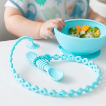 Baby with a patterned shirt uses a blue Silicone Accessory Tether 2-Pack by Bumkins and explores a bright bowl of food on the table.