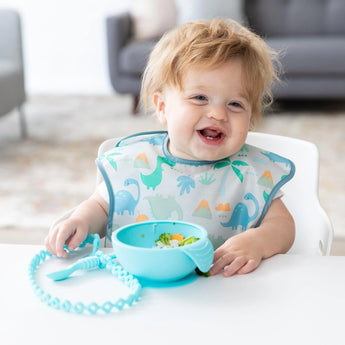 A happy baby in a dinosaur bib uses a Bumkins silicone tether for sensory fun, with a bowl and spoon secured by suction.