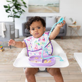 A toddler in a high chair wears a colorful bib, uses Bumkins Silicone Accessory Tether (Blue) for exploration, with food on a suction plate.