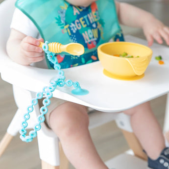 A baby explores a spoon on the Bumkins Blue Silicone Tether. A yellow bowl with a suction cup sits securely on the tray.