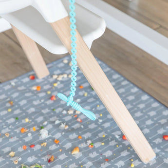 The Bumkins Silicone Accessory Tether 2-Pack in blue holds a spoon, with food scattered on a patterned mat for sensory play.