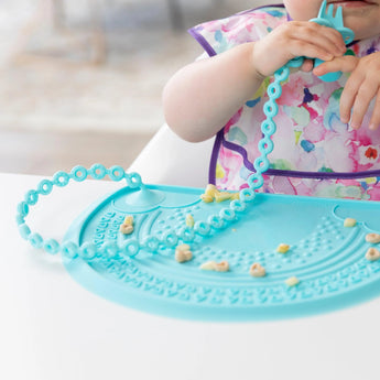 Child in a bib explores a placemat, chewing on a Bumkins Silicone Accessory Tether: Blue, amid small food bits.