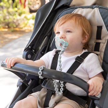 A baby in a stroller with Bumkins Silicone Accessory Tether sits outdoors, wearing a white shirt and shorts, with greenery behind.