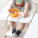 A baby in a gray shirt and sneakers plays by holding a spoon over oatmeal, secured by Bumkins Silicone Accessory Tether, Gray.