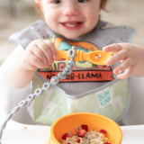 A child with a Silicone Accessory Tether from Bumkins explores sensory delights, using a spoon over cereal in a suction cup bowl.