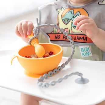 Baby in a high chair explores with Bumkins silicone tethers attached to an orange bowl; bib adorned with playful cartoon animals.