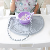 A toddler enjoys yogurt from a purple bowl on a gray mat with a Bumkins Silicone Accessory Tether attached.