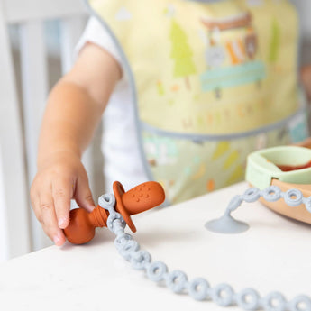 Child holding a brown pacifier with a Bumkins Gray Silicone Accessory Tether, sitting at a white table with a colorful bib and bowl nearby.