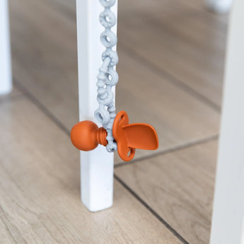 Bumkins Silicone Accessory Tether, Gray, hangs from a clip attached to a white table leg on a wooden floor.