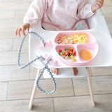 Baby in a high chair with a pink divided plate, enjoying food and exploring with the Silicone Accessory Tether by Bumkins.