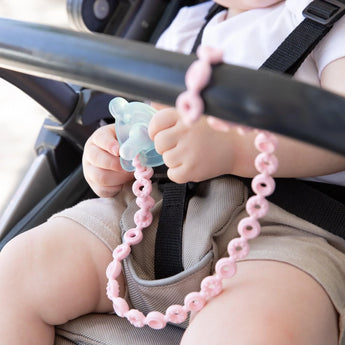 A baby in a stroller, wearing a white shirt and beige shorts, has a Bumkins Pink Silicone Accessory Tether perfect for teething.