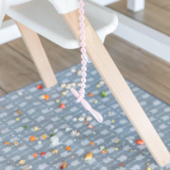 A Silicone Accessory Tether by Bumkins secures a toy to a high chair, above food bits on a wooden floor.