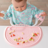 Baby in a rainbow outfit plays with diced fruit on a pink placemat featuring Bumkins Pink Silicone Accessory Tether beaded toy.