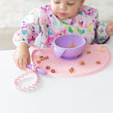 A baby in a bib eats from a purple bowl on a pink mat, holding matching cutlery with Bumkins Silicone Accessory Tether in Pink.