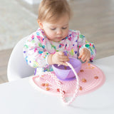 A toddler in a floral bib plays with Bumkins Silicone Accessory Tether, pink bowl, on a pink placemat at the white table.