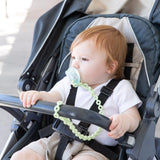 A baby with a pacifier in a stroller, wearing a white shirt and beige shorts, uses Bumkins Silicone Accessory Tether in Sage.