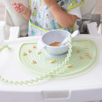 A child in a high chair enjoys a meal with Bumkins Silicone Accessory Tether on a green placemat, secured by sleek suction.