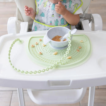 A baby in a high chair enjoys food on a silicone mat with a Bumkins sage silicone tether and teething necklace draped on the tray.