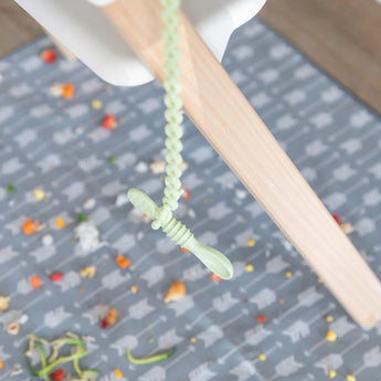 Sage Silicone Accessory Tether from Bumkins secures the spoon to the high chair leg, food rests on a patterned mat below.