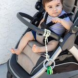 A baby in a stroller joyfully gazes at the camera with a toy on a Bumkins Sage Silicone Accessory Tether dangling from their tray.