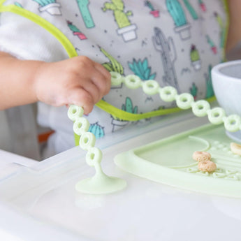 A baby in a high chair plays with cereal on a Bumkins Silicone Accessory Tether in Sage, wearing a cactus-patterned bib.