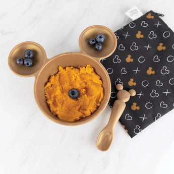 Silicone bowl set by Bumkins with orange puree and blueberries on black cloth; perfect for your little ones first feeding. Minnie Mouse Gold design.