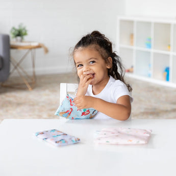 A child beams at a table with a colorful book and Bumkins Princess Magic, Ariel, and Jasmine Reusable Snack Bags in the bright room.