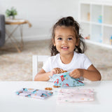 Young girl grins at the table, holding a Bumkins Princess Magic Snack Bag in the bright room with white shelves.