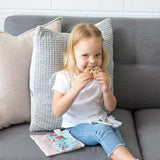 A blonde girl in a white shirt and jeans smiles on a gray couch, enjoying cookies from her Bumkins Princess Magic Snack Bag.