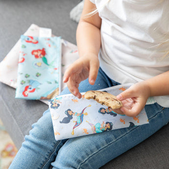 Child in jeans on grey couch holding half-eaten cookie with colorful Bumkins Princess Magic, Ariel, and Jasmine snack bags.