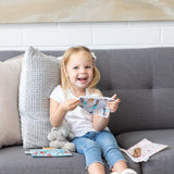 A smiling young girl on a gray sofa holds a patterned pouch with Bumkins Disney Princess Snack Bags and a stuffed animal beside her.