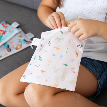 Child on couch, opening Bumkins reusable snack bag from the 3-Pack: Princess Magic, Ariel, and Jasmine.