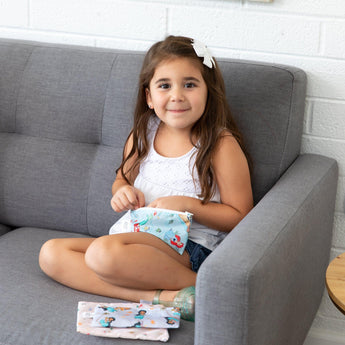 A girl with long hair sits on a gray sofa, holding a Bumkins Reusable Snack Bag: Princess Magic. Two more bags are beside her.